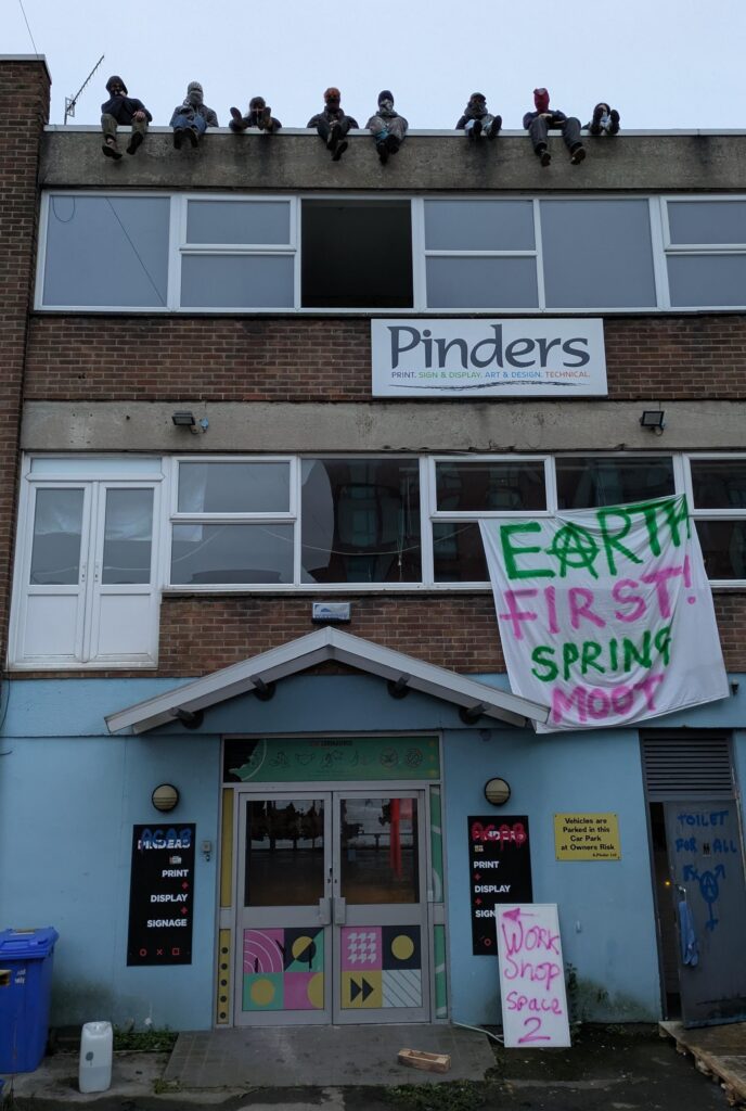 A picture of a 3 storey squat of an old printing shop. there is a banner hanging out one window reading Earth First! spring moot. On the ground floor there's a large spray painted sign reading workshop 2. Acab has been painted on the old logos. There are 8 masked figures sitting on the edge of the roof at the top of the photo