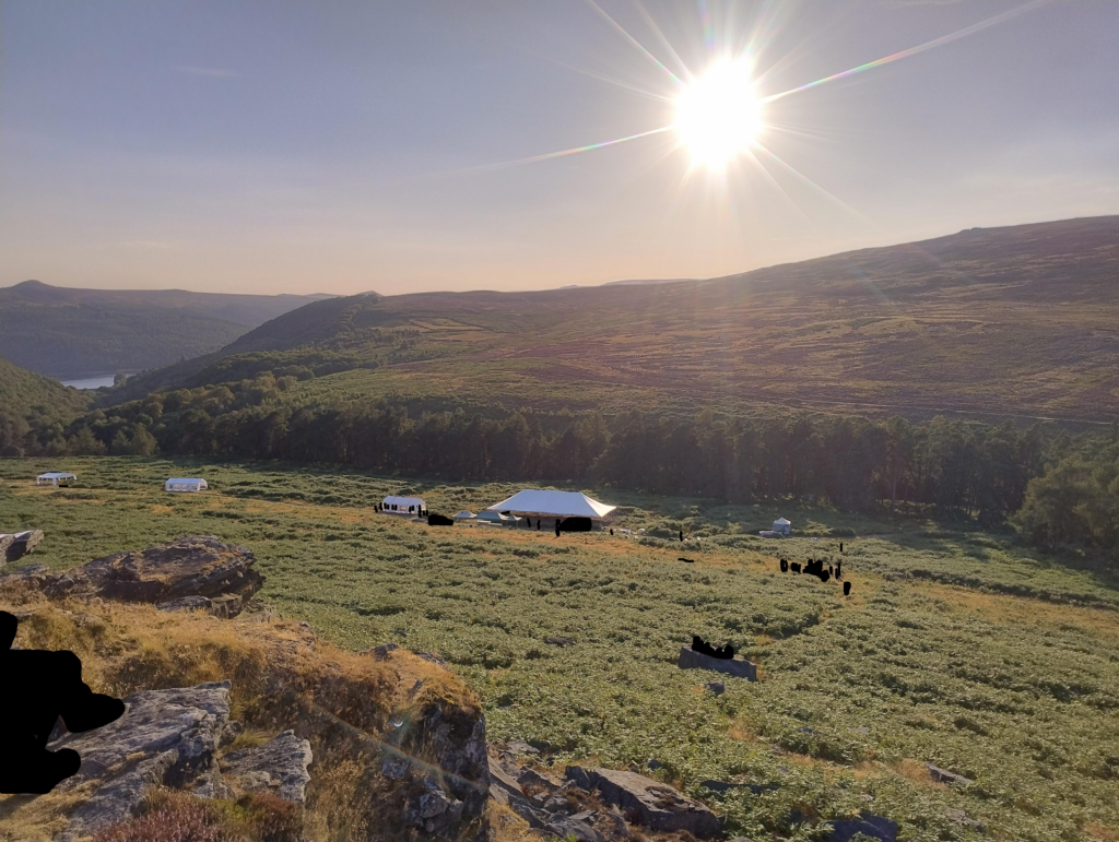 four large marquees on a beautiful hillside are shown. the peak district heather hills are on the horizon. the evening sun light is dimmed by the eclipse.